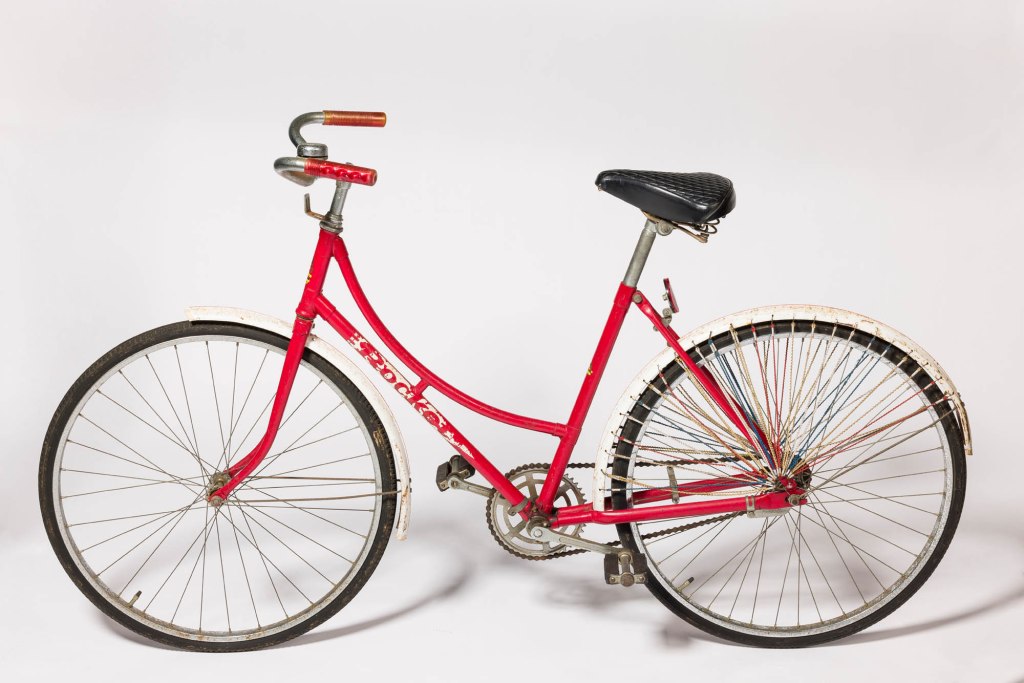 A vintage 'Rocket' bicycle with a red steel frame, single-speed mechanism, and classic styling, featuring prominent wheels and handlebar grips.