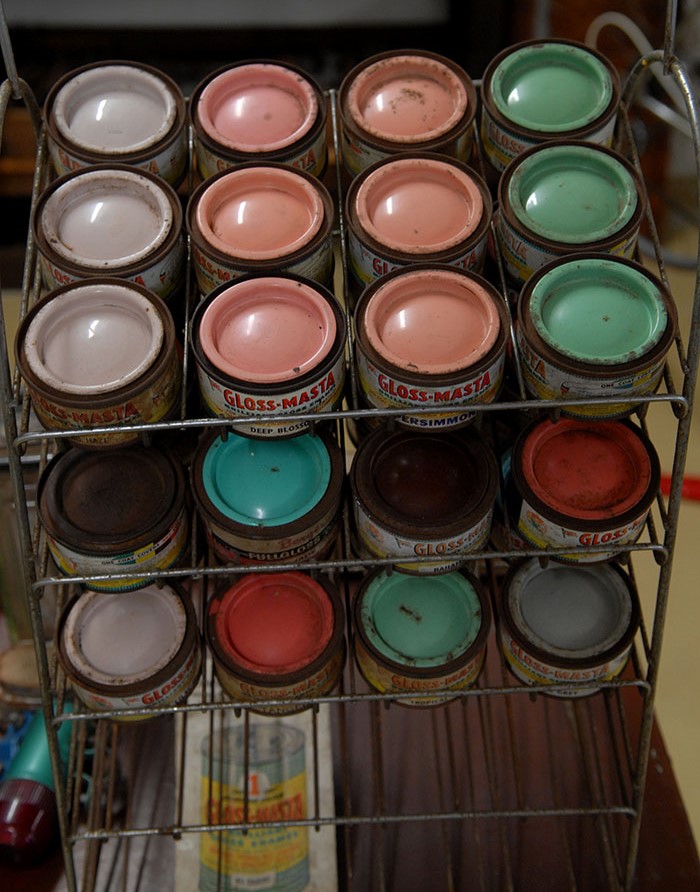 A metal rack displaying various cans of Gloss-Masta paint, showcasing a selection of colors including pink, green, and cream, some with dust on the lids.