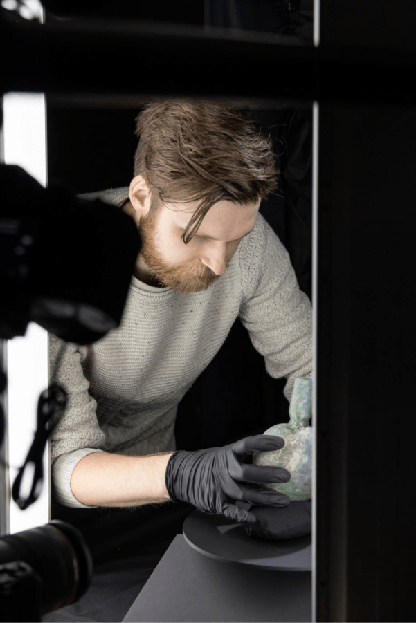 A man in a gray sweater and black gloves carefully examines an ancient artifact in a dimly lit environment, focused on detailing and preservation.