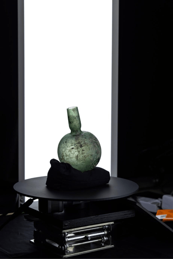 A green glass bottle on a black cloth, displayed on a rotating stand against a bright white background.