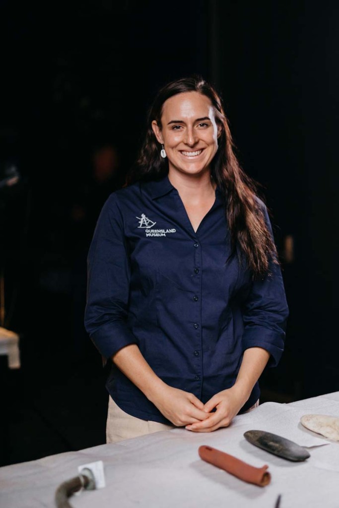 A smiling woman wearing a blue shirt with the Queensland Museum logo, standing in front of a table with various tools or artifacts.